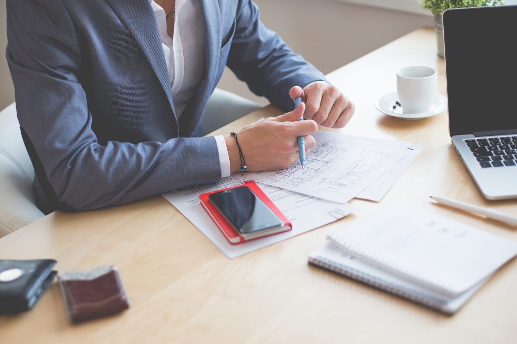 Person in suit writing at a desk, laptop, phone, notebook, coffee cup, and wallet visible.