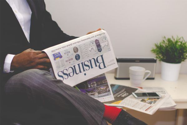 Person in suit reading a business newspaper; desk with plant, speaker, phone, and mug.