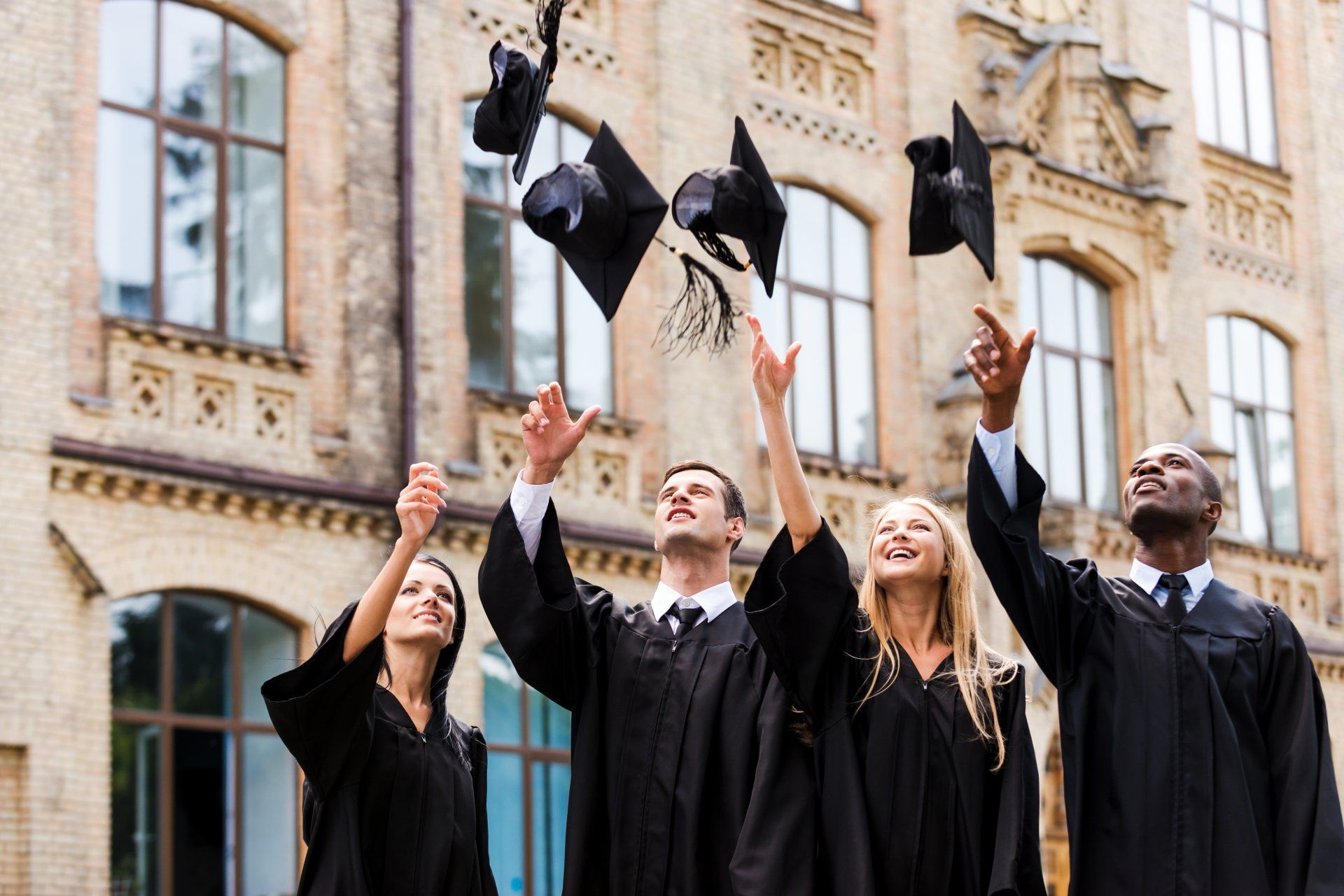 Graduates in black gowns toss mortarboards in air outside a brick building.