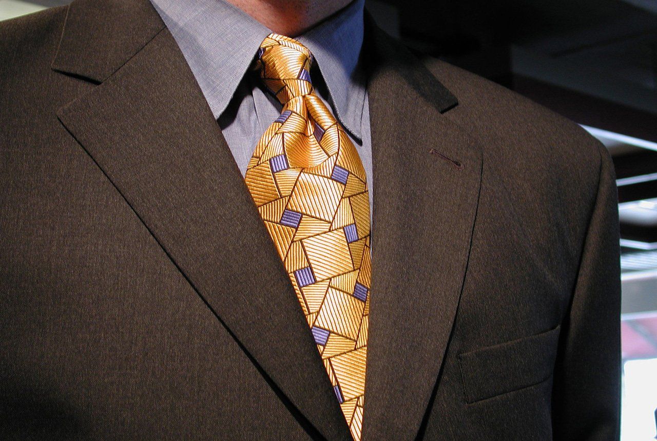 Man in dark suit, light blue shirt, and gold geometric-patterned tie.