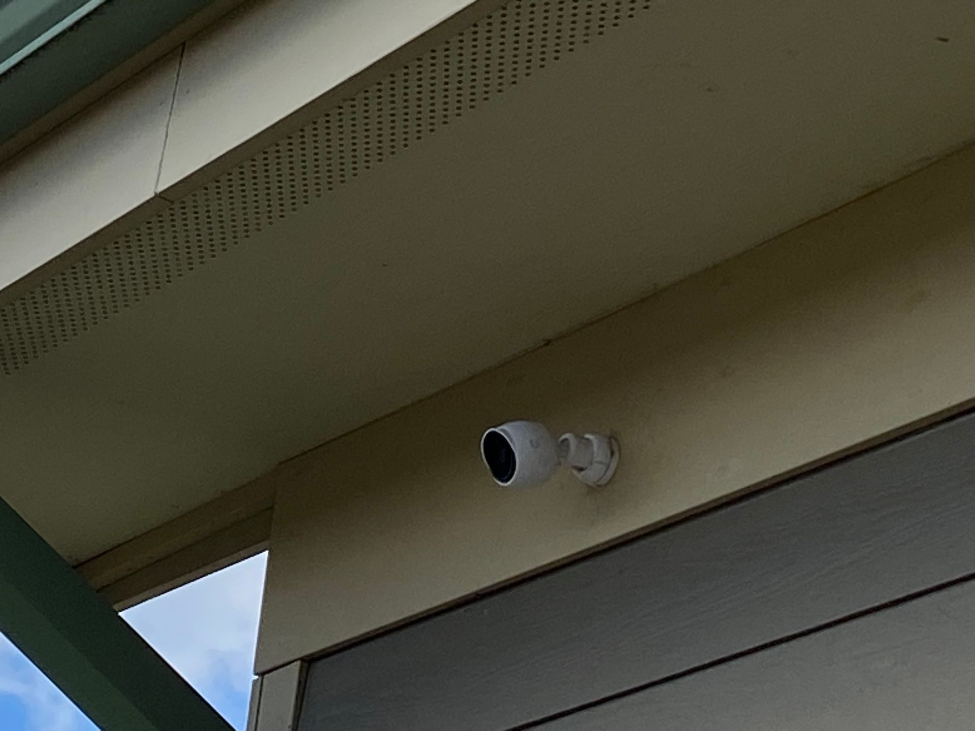 A white outdoor security camera mounted on the cream-colored wall of a house under a roof overhang.