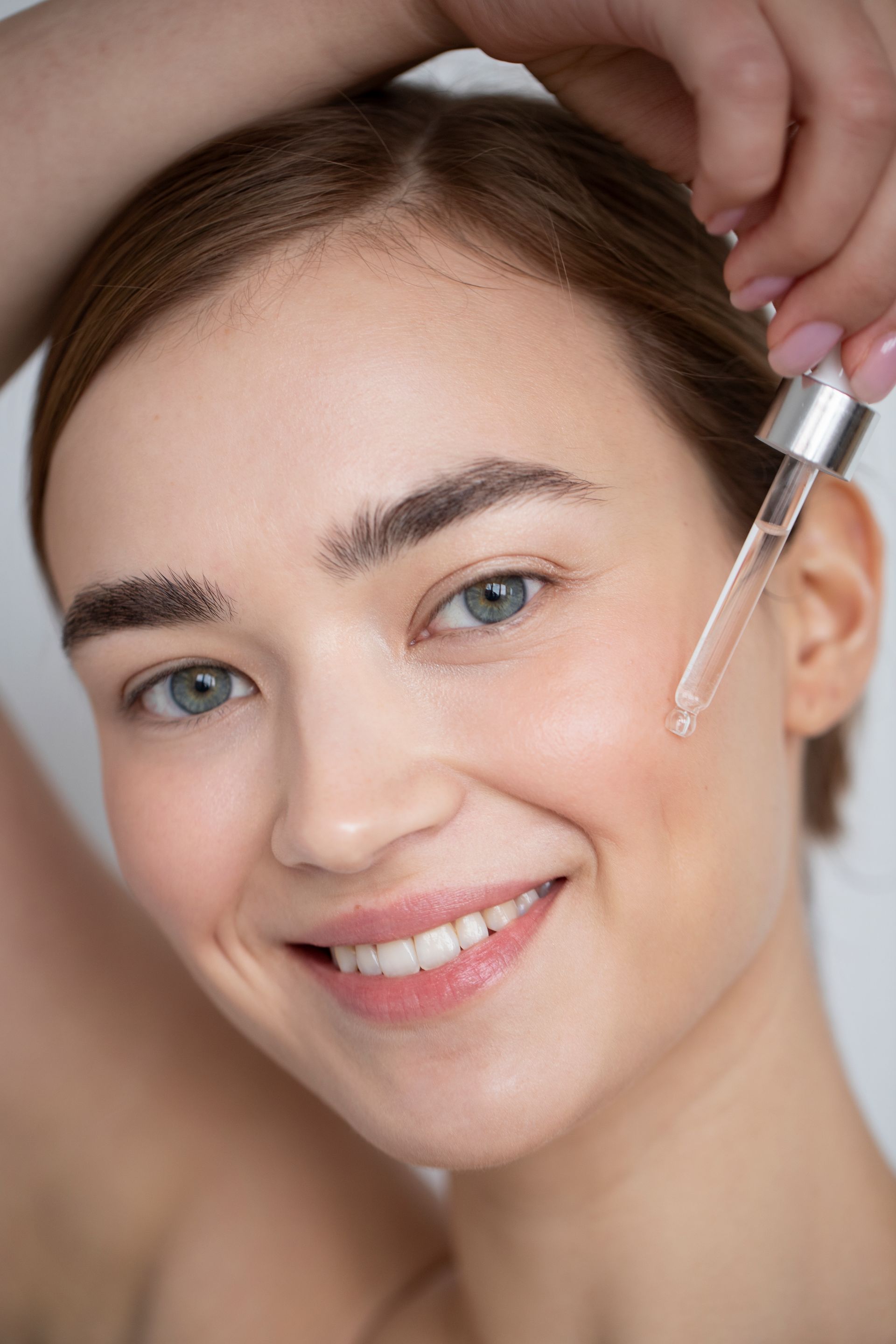 A woman is smiling while applying a serum to her face.