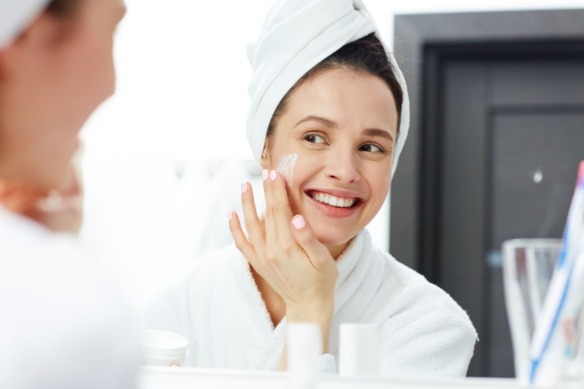 A woman with a towel wrapped around her head is applying cream to her face in front of a mirror.