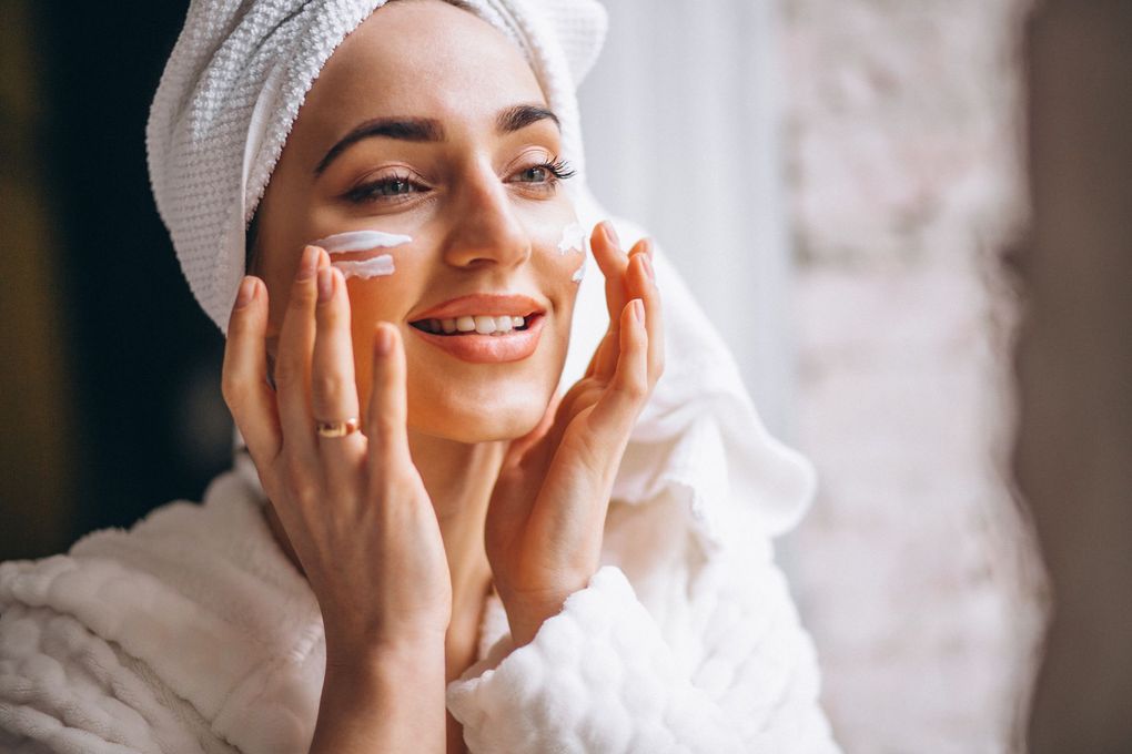 A woman with a towel wrapped around her head is applying lotion to her face.