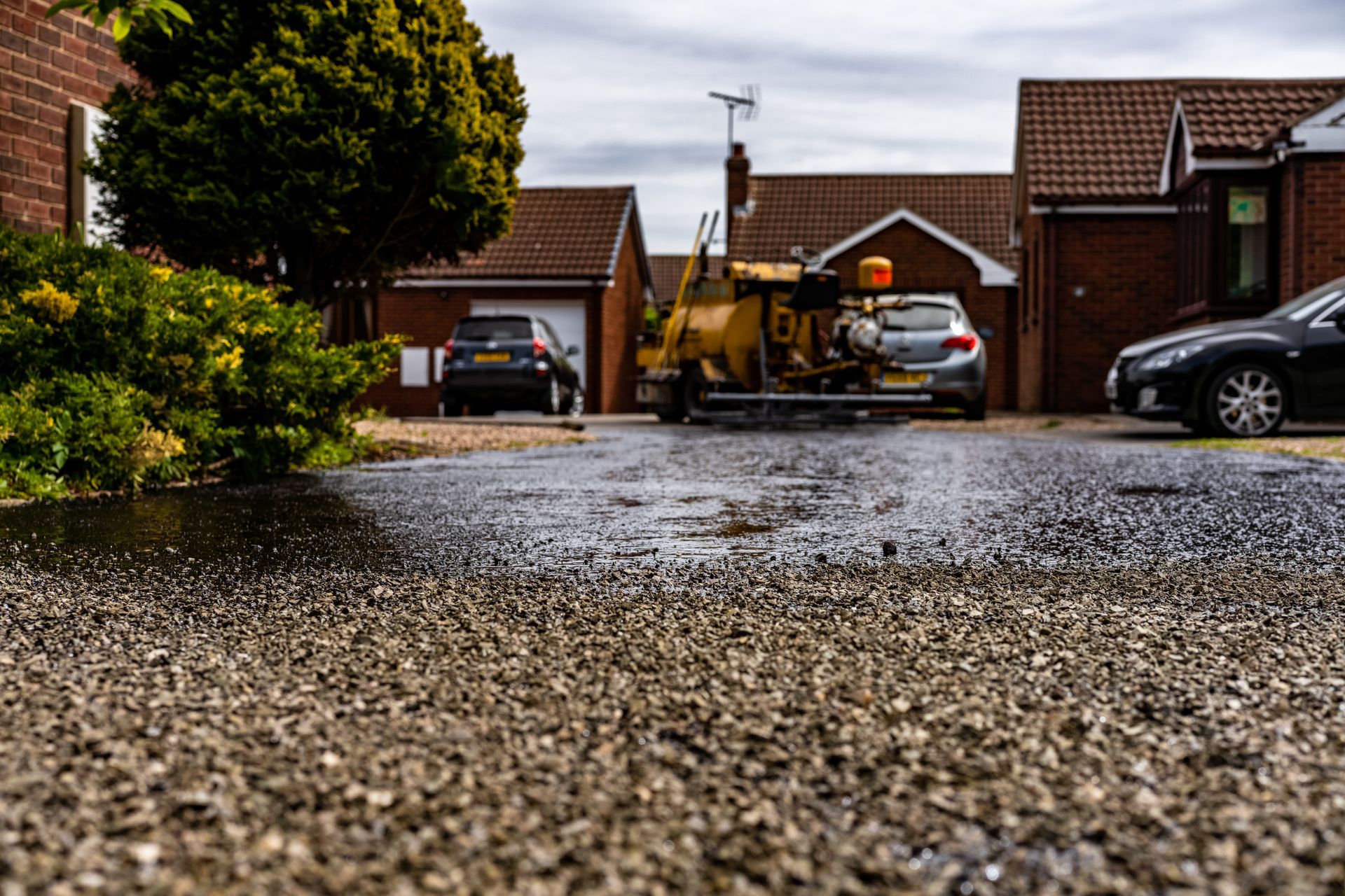 Rain-soaked gravel driveway with suburban houses, parked cars, and a small yellow construction vehicle