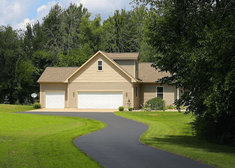 Suburban beige house with three-car garage, curved driveway, and green lawn surrounded by trees