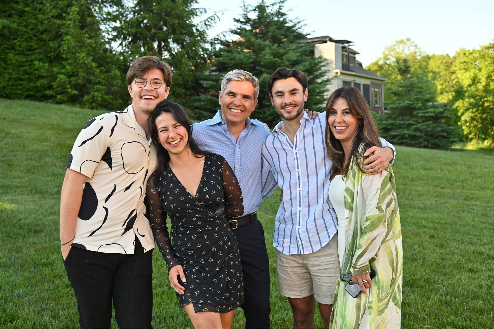 Family of five smiling, standing on a grassy lawn with a house and trees in the background.