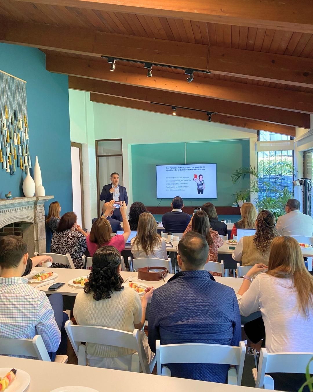 A man presents to an audience. Group sits at tables, looking toward him. Presentation displayed on screen.