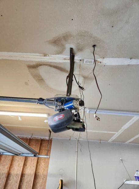 Garage ceiling with a blue and black garage door opener hanging from exposed wiring and tracks