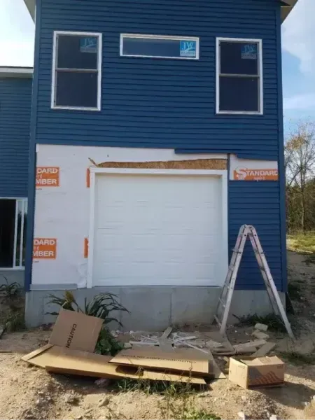 Blue house exterior under construction with white garage door, ladder, and building materials in front