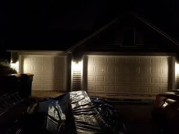 Dark suburban house at night with lit garage doors and a tarp-covered object in the driveway