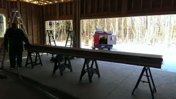 Long beam resting on sawhorses inside a workshop, with a person standing nearby and a wooded area outside.