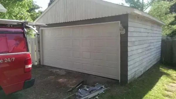 Detached white garage with a closed beige door, seen beside a red vehicle in a grassy yard