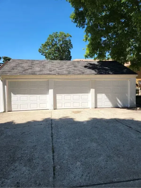 Three-car garage with white doors and a dark shingled roof, viewed from a sunlit driveway.