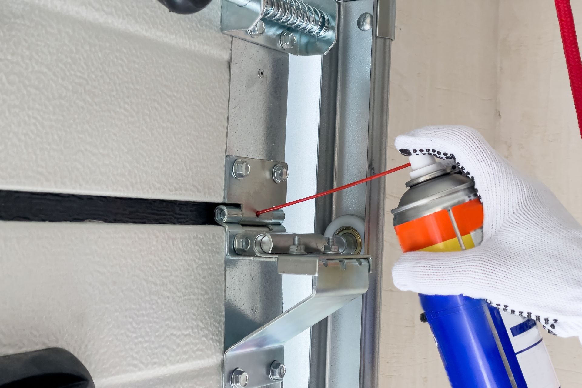 Gloved hand spraying lubricant on a metal garage door hinge and track.