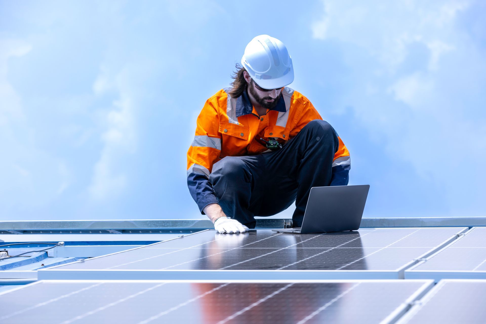 View of a solar panel technician with safety gear checking a panel against blue sky. View of a solar panel technician with safety gear checking a panel against blue sky.