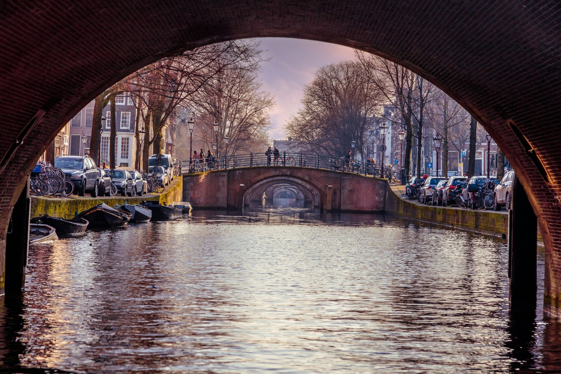 Canal view in Amsterdam, framed by a bridge arch, with parked cars, trees, and brick bridge.