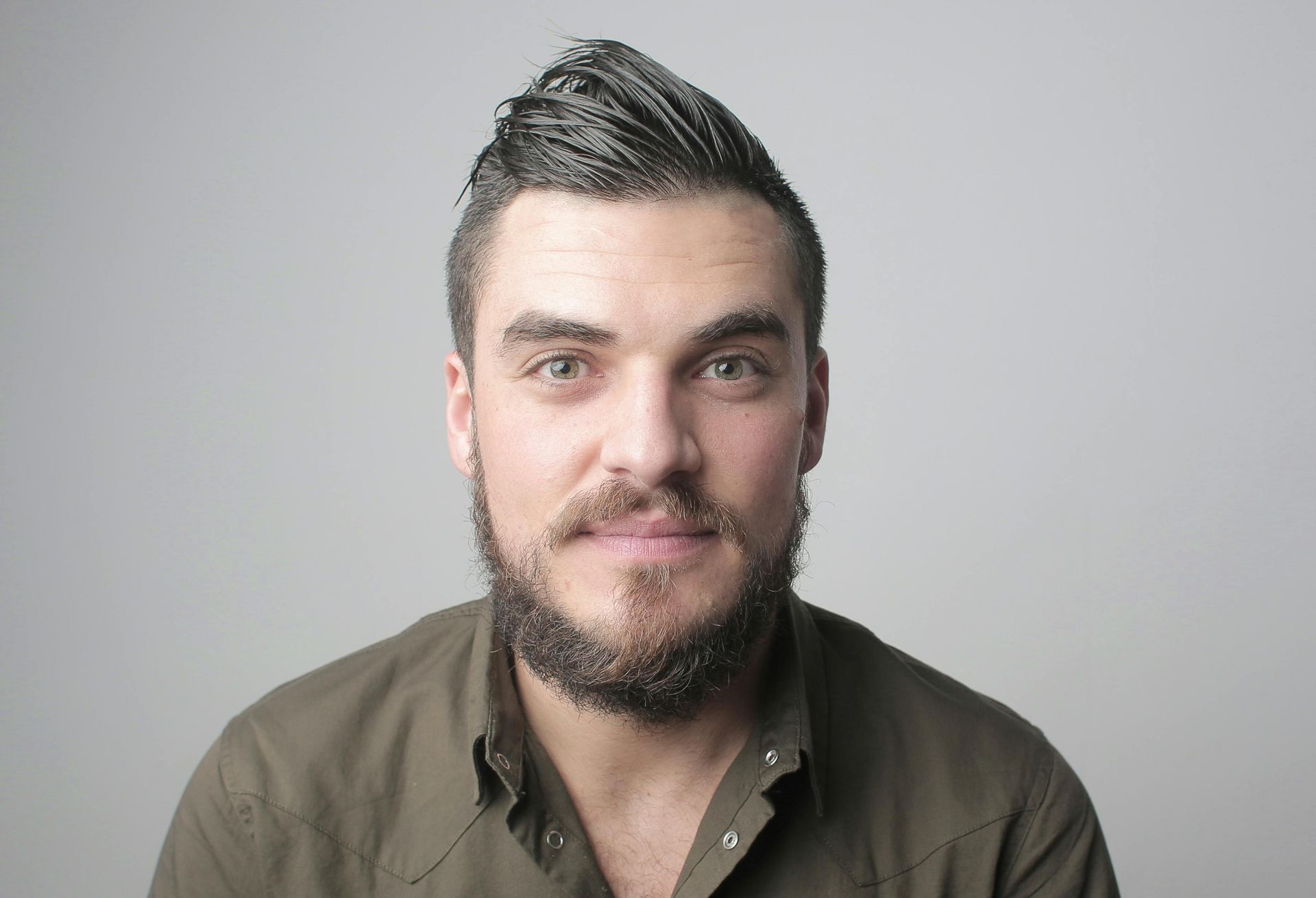 Man with a beard and mohawk looking at the camera, wearing a green shirt, against a grey backdrop.