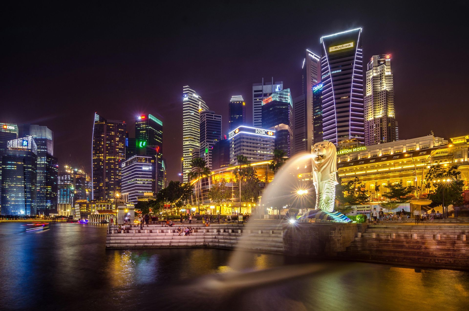 Night view of Singapore skyline with illuminated skyscrapers and the Merlion statue spouting water.