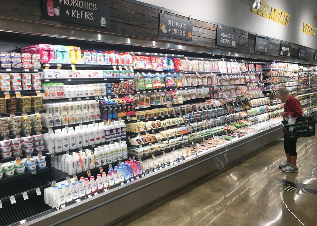 Person browsing dairy aisle in a grocery store. Refrigerated shelves stocked with yogurt and kefir, with overhead signage.
