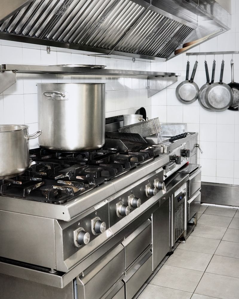 Commercial kitchen with stainless steel stove, pots, and hanging pans under a ventilation hood.
