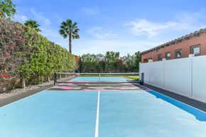 Blue outdoor sports court with tennis net, palm trees, and a building beside a white fence