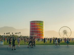 Crowd at an outdoor festival near a colorful cylindrical tower and Ferris wheel at sunset.