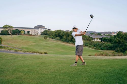 Golfer mid-swing on a green course with trees and buildings in the background