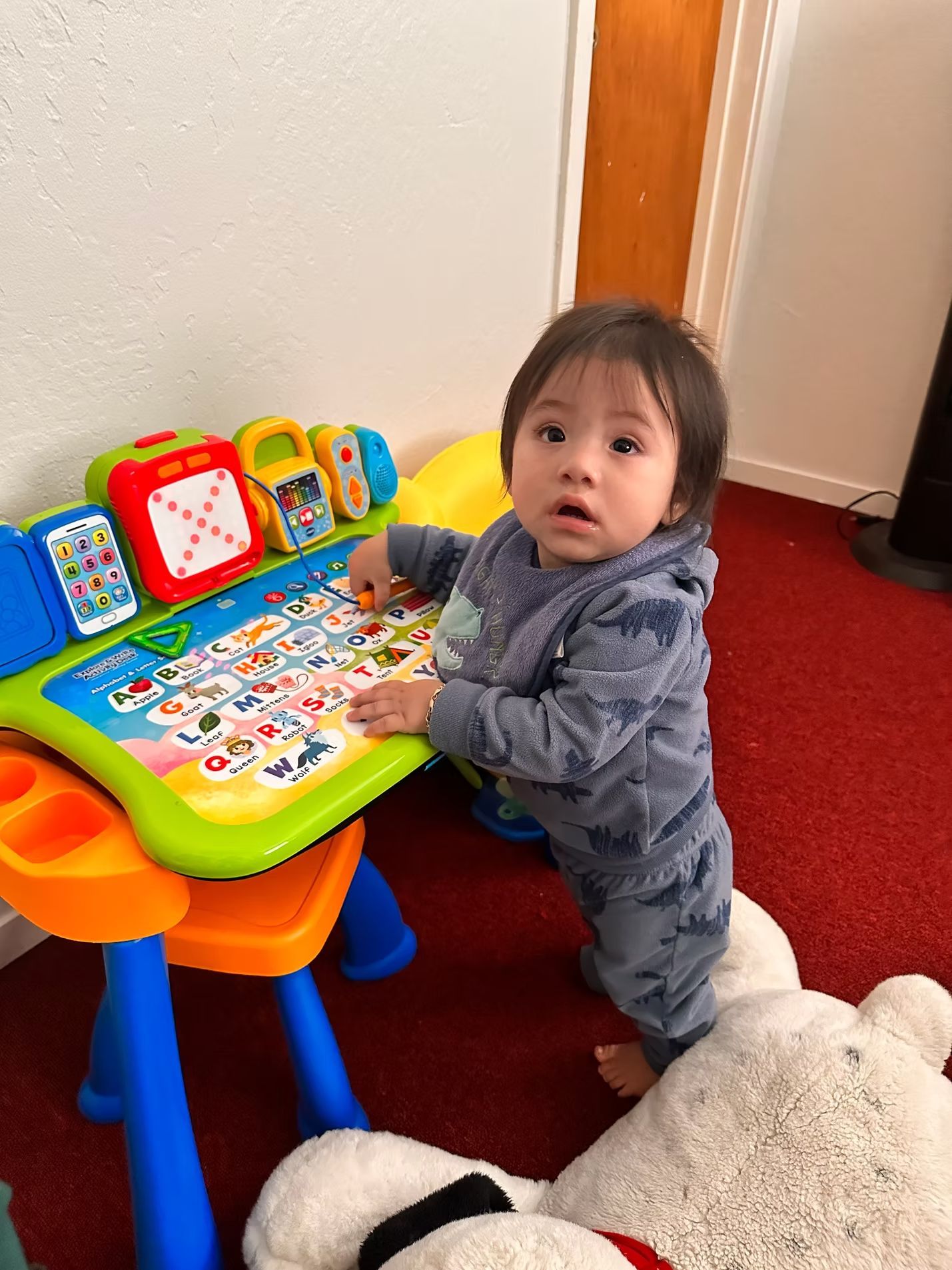 Toddler standing by a colorful activity table, looking up indoors on a red carpet.