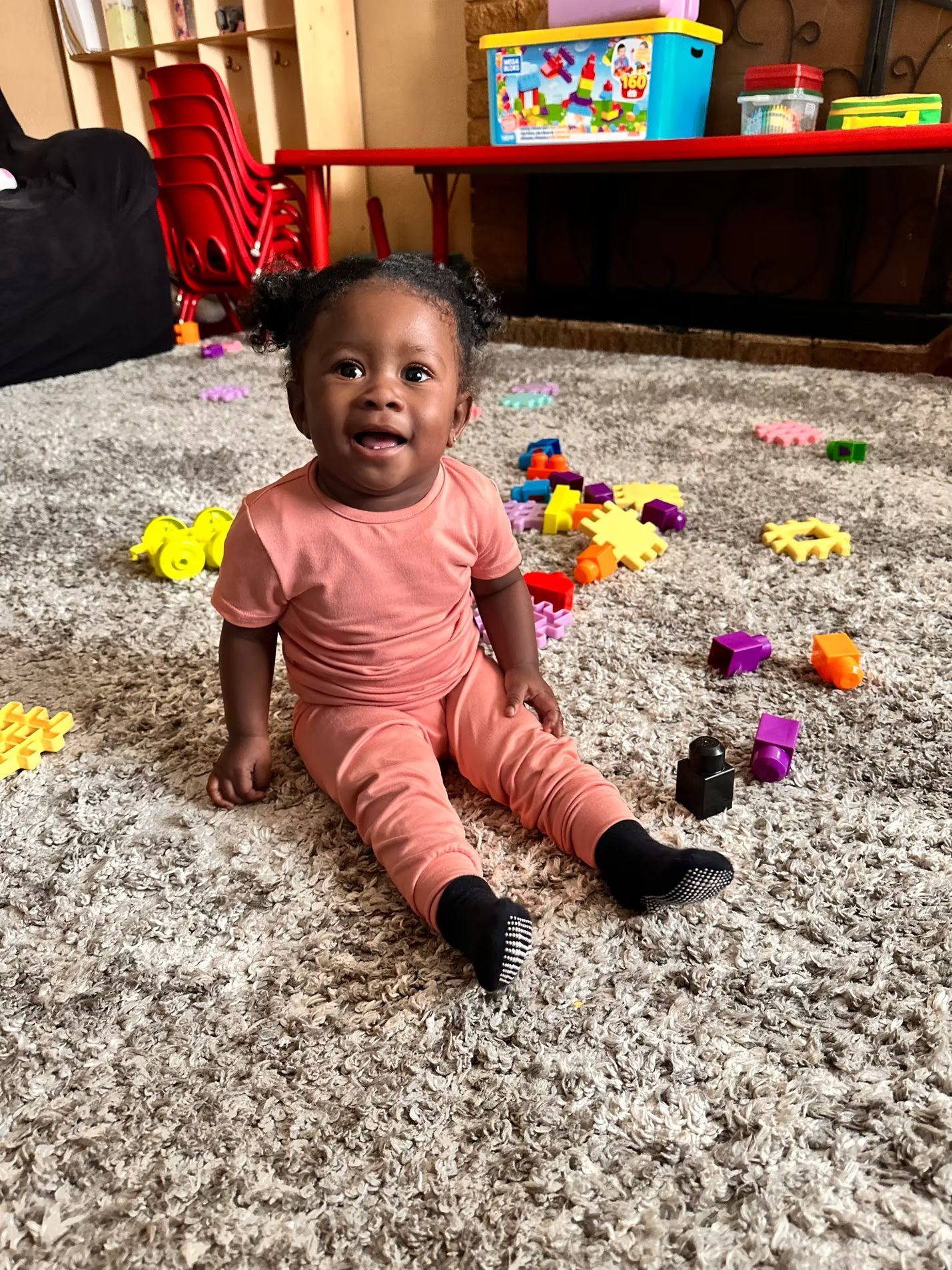 Child sitting on a carpet, smiling amid colorful toys in a living room.
