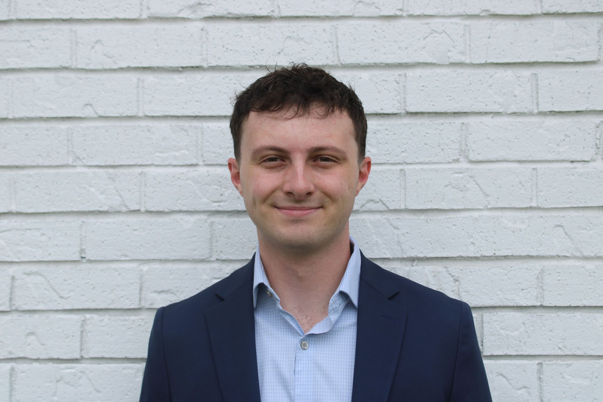 Man in a navy blazer and light blue shirt standing against a white brick wall, smiling at the camera