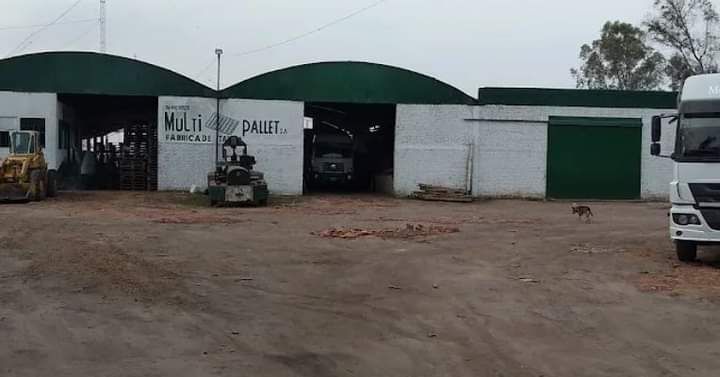 Patio industrial con cobertizos de techo verde, una carretilla elevadora y un camión blanco en un terreno de tierra.