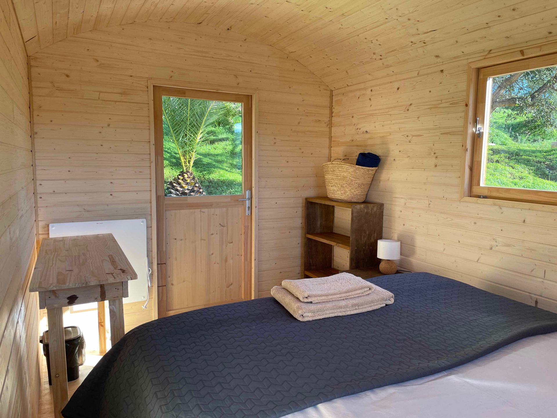 Small wooden cabin bedroom with a bed, shelves, and two windows overlooking greenery.