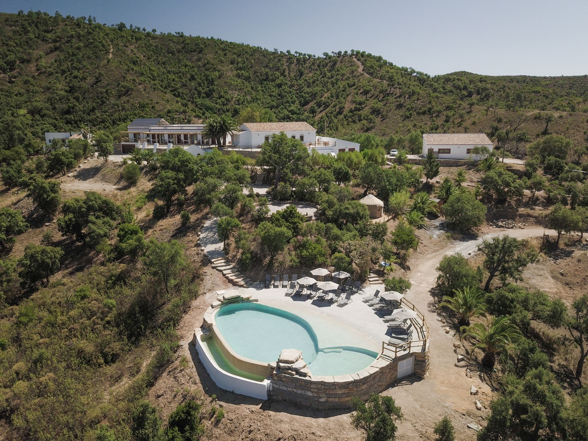 Aerial view of a hillside resort with a turquoise pool and scattered white buildings among dry green trees.
