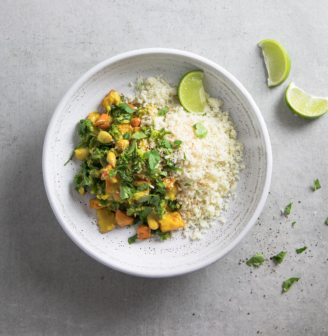 Rice bowl with colorful vegetable curry, garnished with cilantro and lime wedges on a gray surface