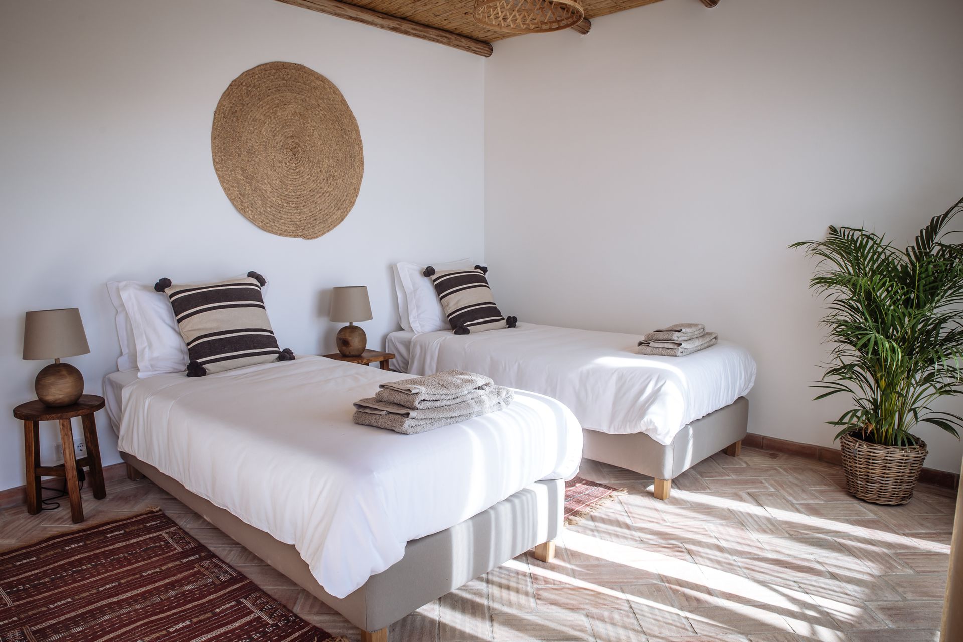 Bright bedroom with two white twin beds, woven wall decor, and sunlight on a rug