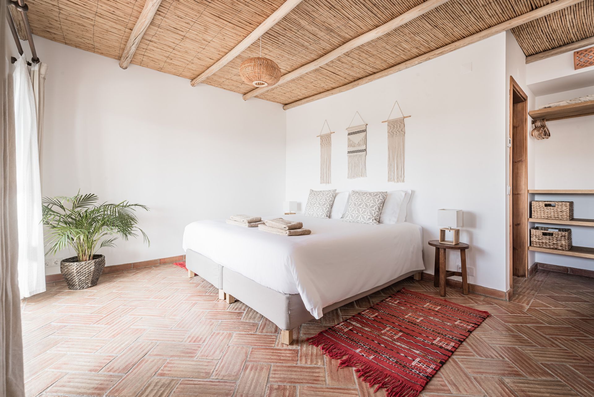 Bright bedroom with white bed, woven ceiling, brick floor, and red rug near a windowed wall