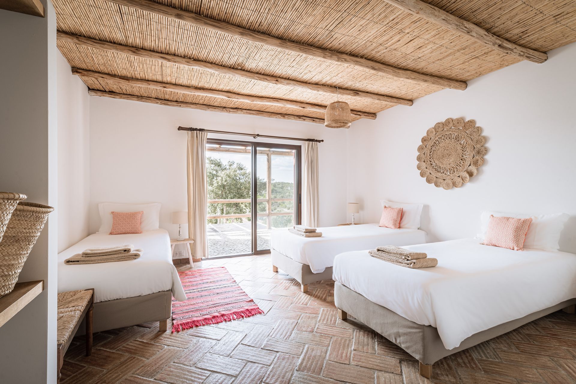 Bright bedroom with two white beds, thatched ceiling, woven decor, and sliding doors to a balcony.