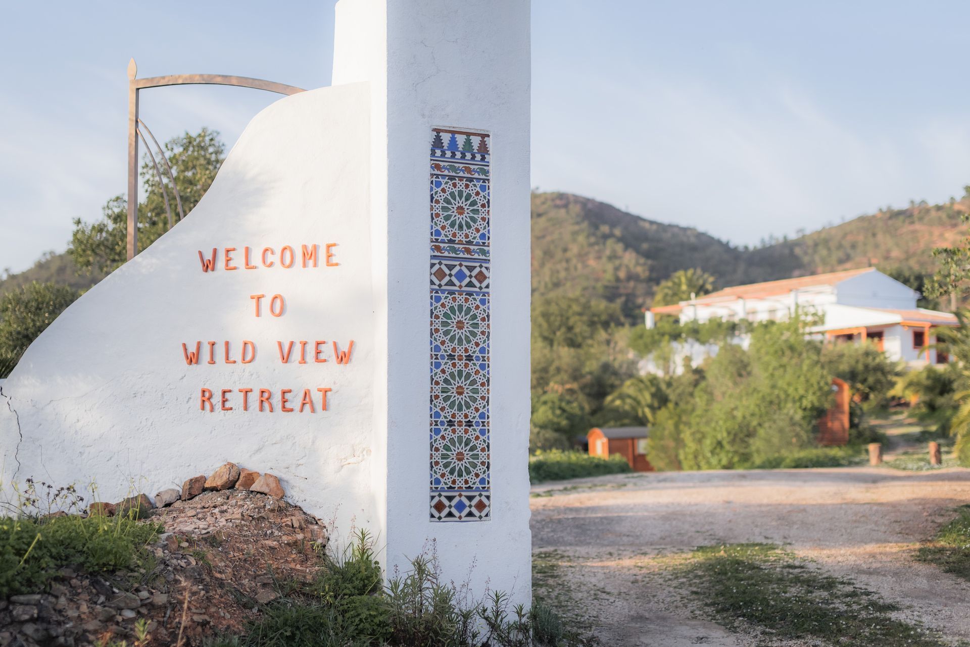 Entrance sign for Wild View Retreat with white wall, mosaic pillar, and hillside buildings in the background