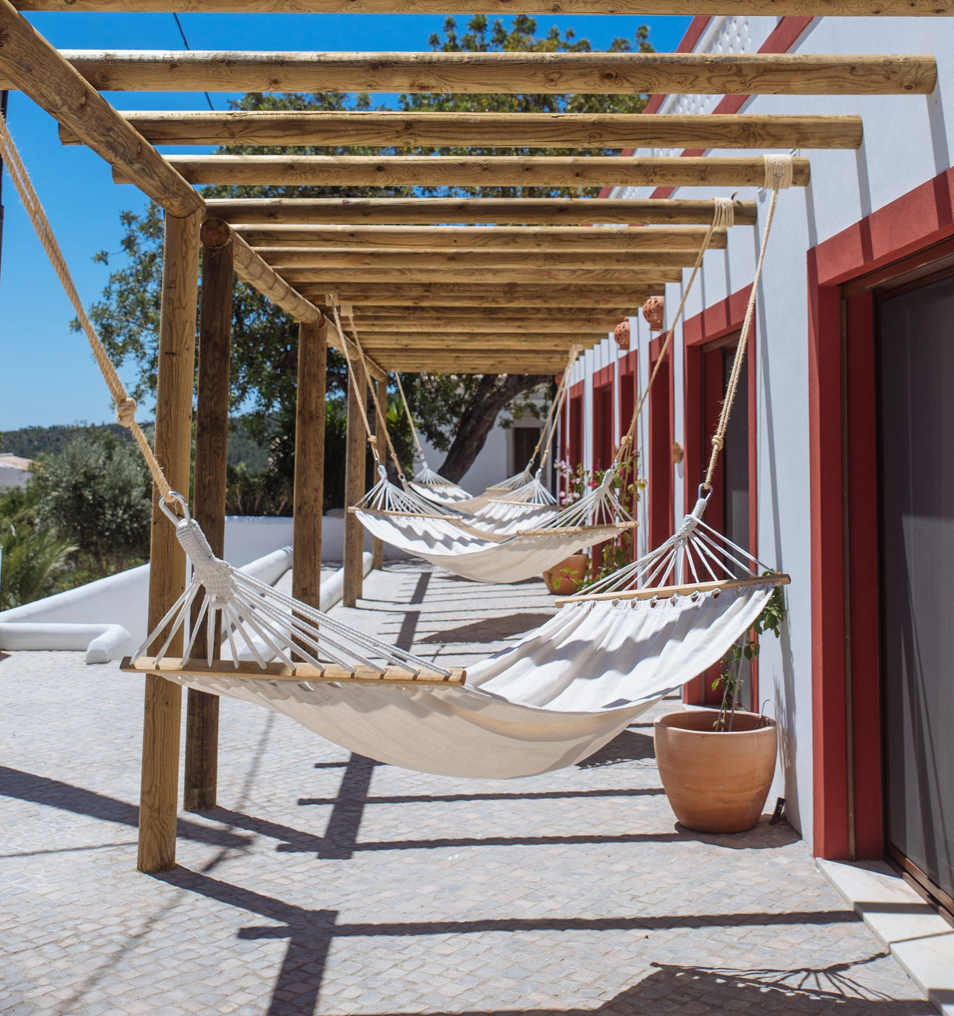 Covered terrace with wooden pergola and hanging hammocks beside a red building, overlooking a scenic view