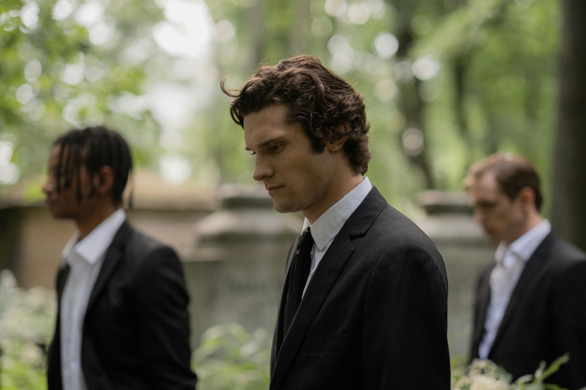 Three men in dark suits standing outdoors, with the central man looking down pensively.