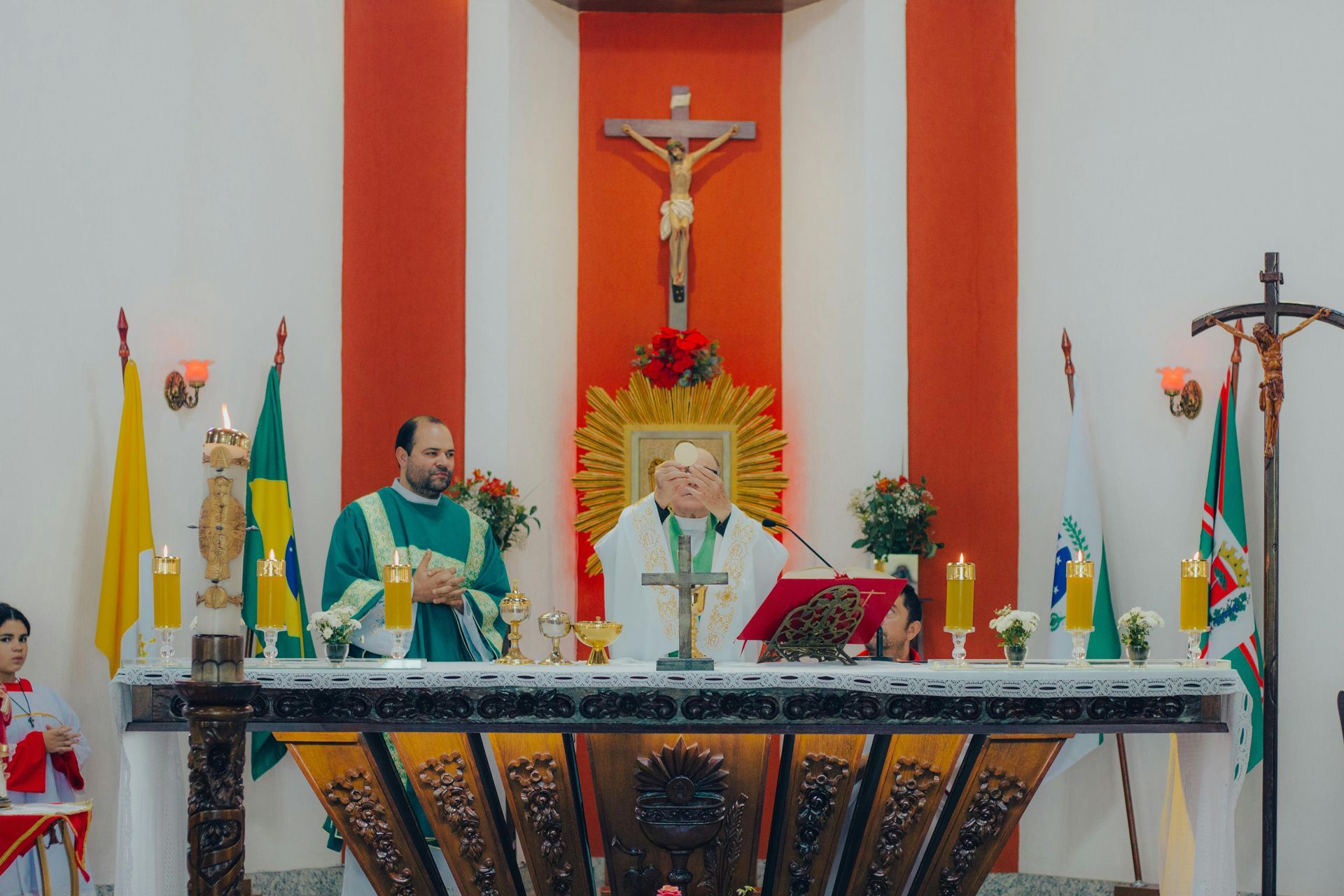 Catholic altar with crucifix, priest in green vestments, and floral arrangements in a church sanctuary