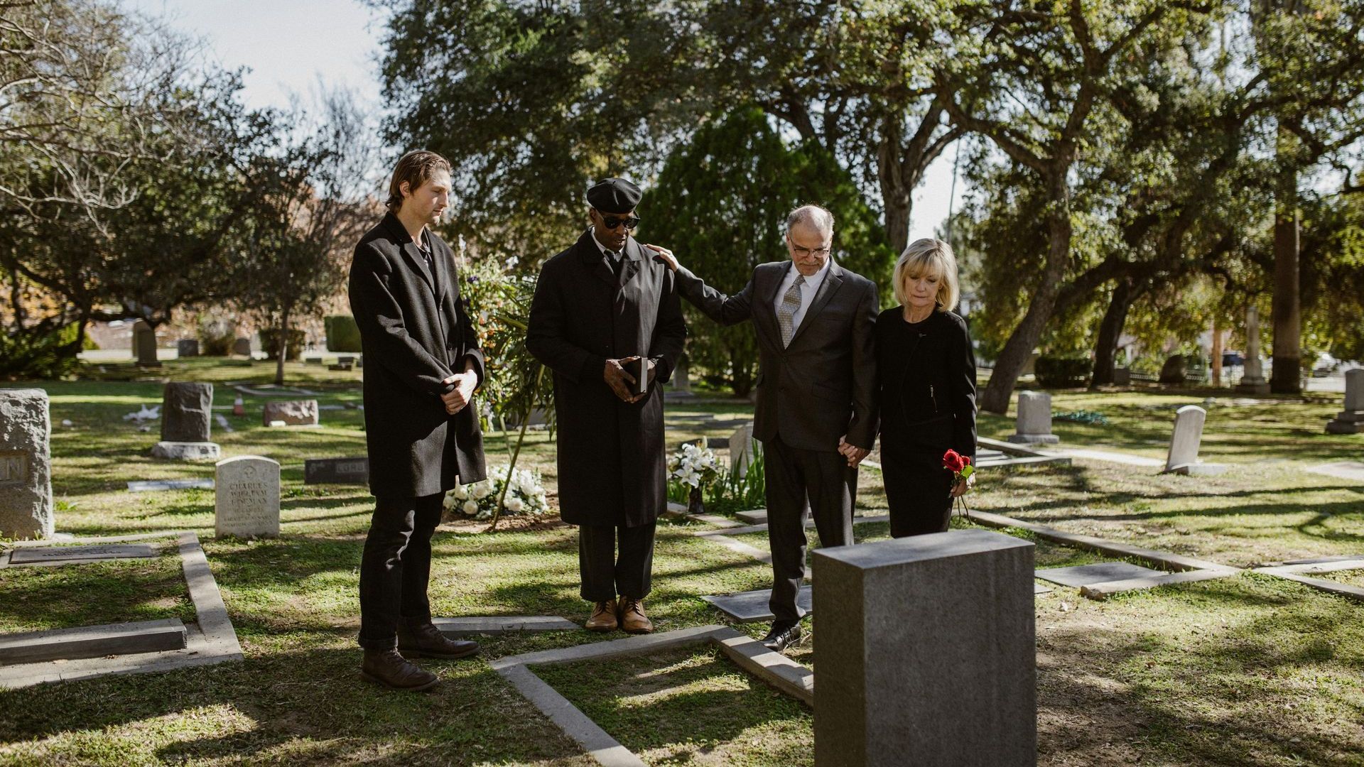 Four people stand among gravestones in a sunny cemetery during a solemn visit.