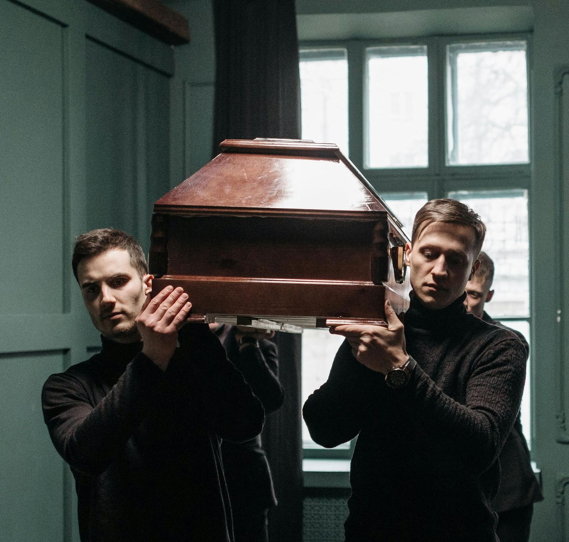 Two men in black carry a wooden coffin indoors by a window