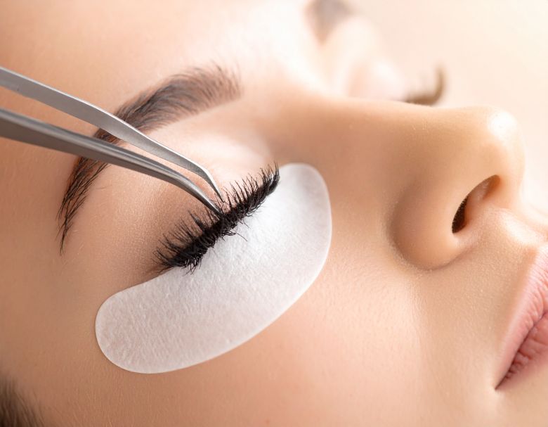 Person receiving eyelash extensions. Tweezers hold a lash near the eye with a pad beneath.