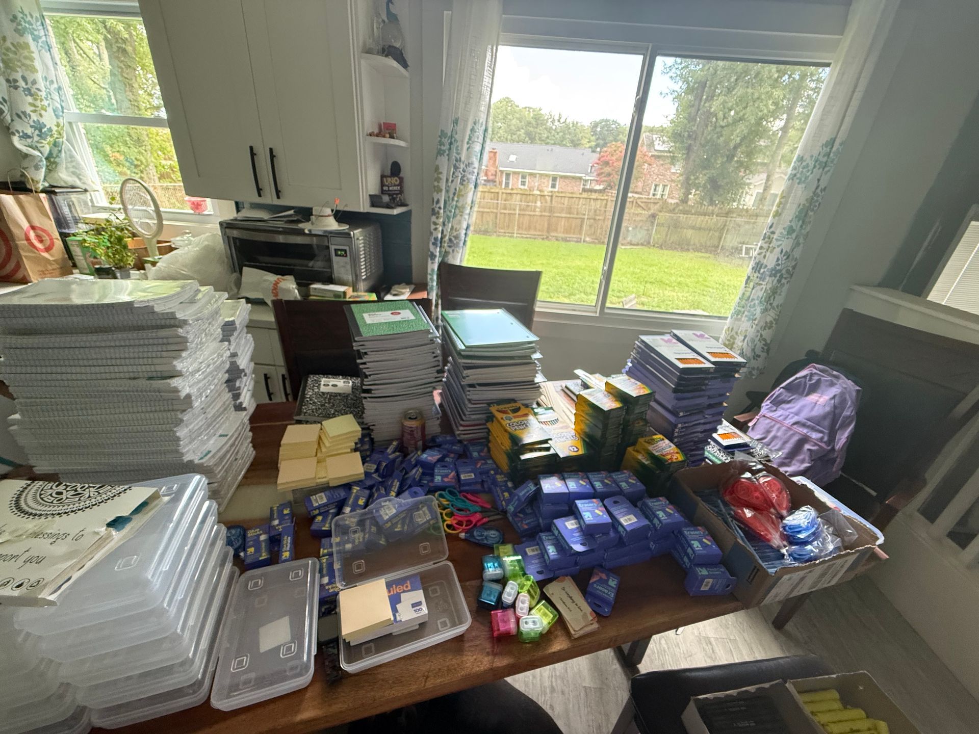 Table covered with stacked supplies, plastic containers, and packaged goods in a bright kitchen.