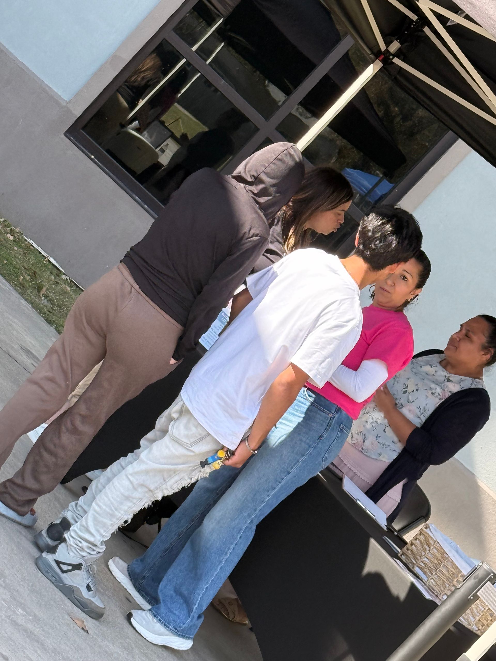 People gathered around a table outdoors beside a building, leaning in to look at something.