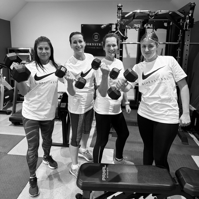 Four women in a gym, holding dumbbells, wearing white shirts with logos, smiling.