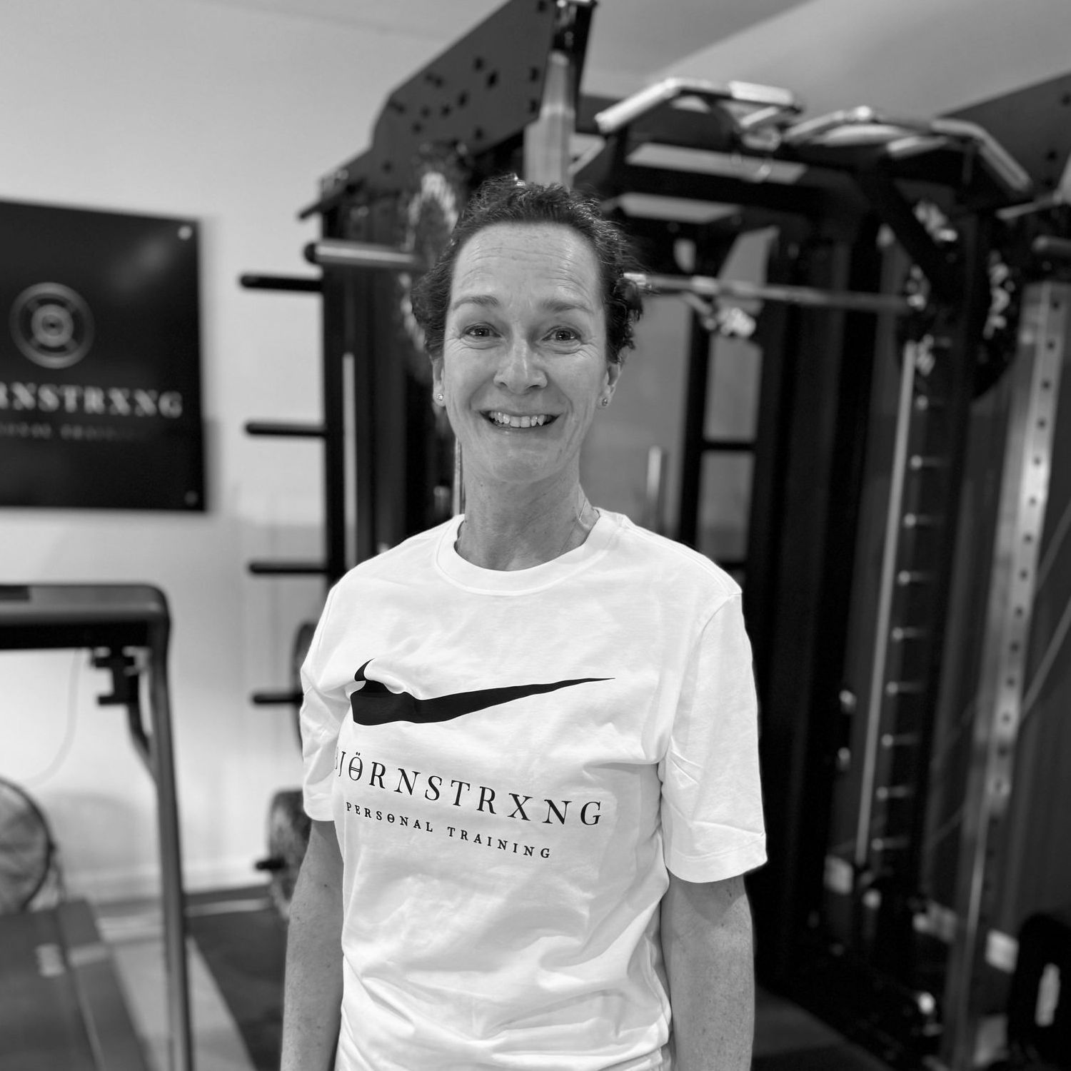 Woman in white t-shirt with logo, smiling in a gym setting, with exercise equipment in the background.