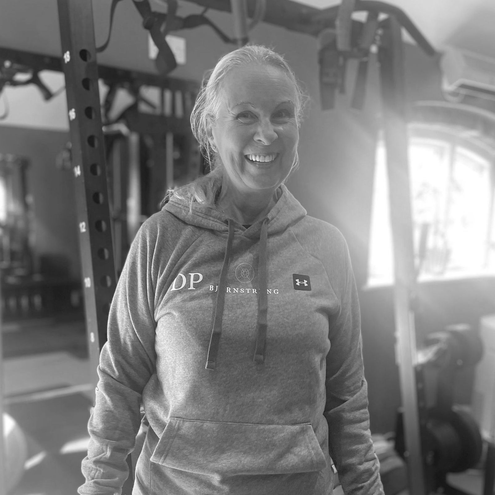 Woman in gray hoodie smiles in a home gym setting, with exercise equipment in the background.